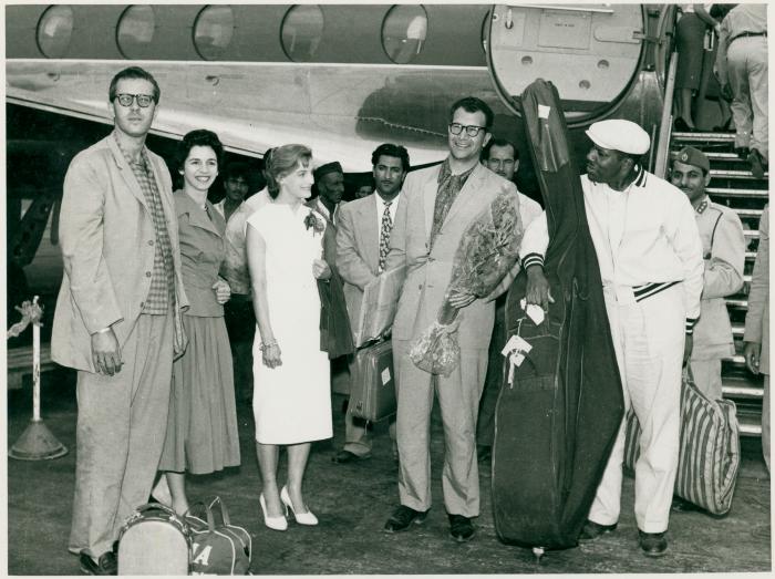 Dave Brubeck (holding bouquet), Joe Morello, Eugene Wright with his bass and assorted embassy personnel in front of airplane (Baghdad, Iraq)